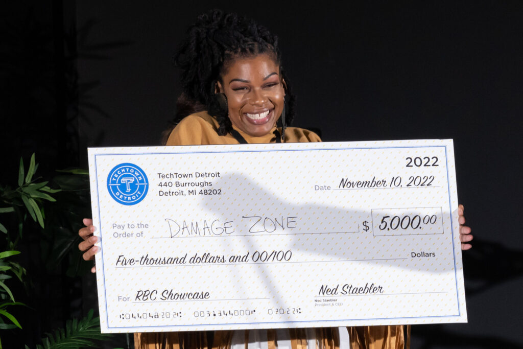 A woman smiles and holds a check for $5,000 from TechTown Detroit for her Detroit-based rage room and axe-throwing business, The Damage Zone.