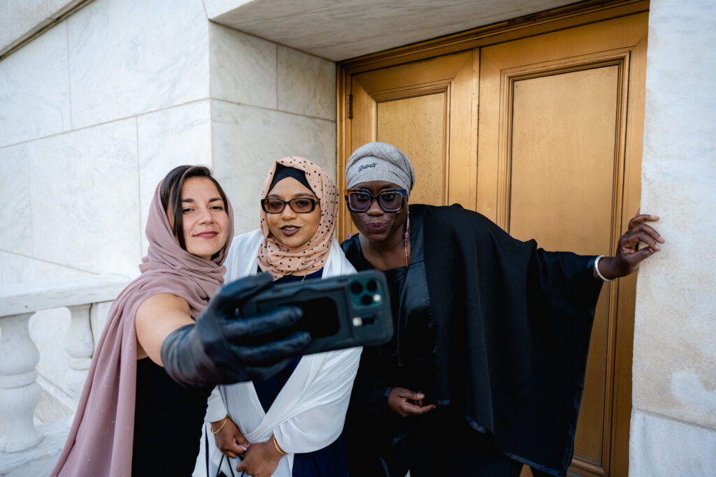 Three women pose to take a selfie outdoors in front of a marble wall and a brown doorway. The woman on the left is holidng the phone. She has on a mauve hijab, a black top and black elbow-length gloves. The woman in the center has on peach-colored hijab with black polka dots, black sunglasses and a white long sleeve shawl with a black top underneath. The woman on the right wears a gray turban with the word "Detroit" embroidered in the center, black sunglasses, a black top and a black jacket.