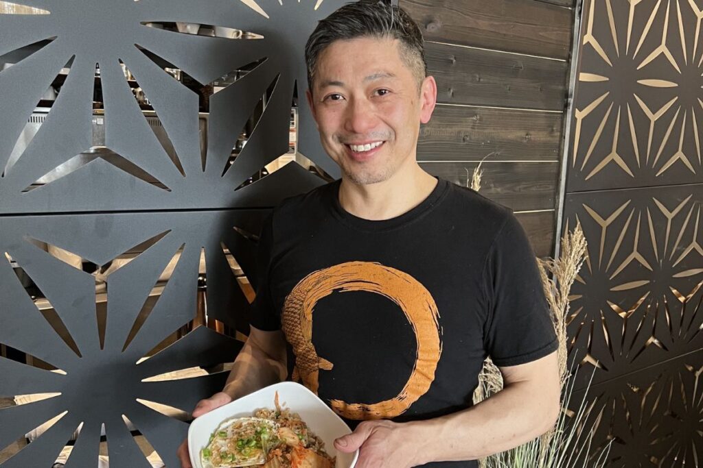 A man wearing a black shirt smiles and holds a bowl of food