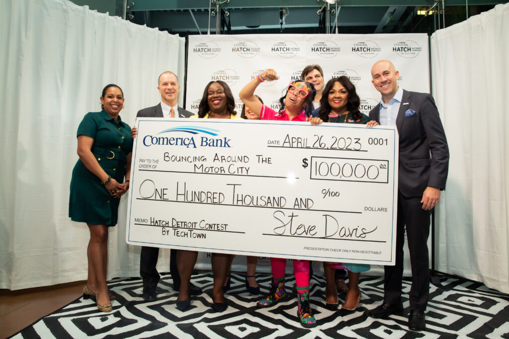Two rows of people stand and pose for a photo. They are standing on a white-and-black patterned rug and in front of a white curtain and step-and-repeat with the Comerica Hatch Detroit Contest by TechTown logo on it. The four people standing in the front are holding a large check from Comerica Bank for $100,000, that is going to the business Bouncing Around the Motor City. 