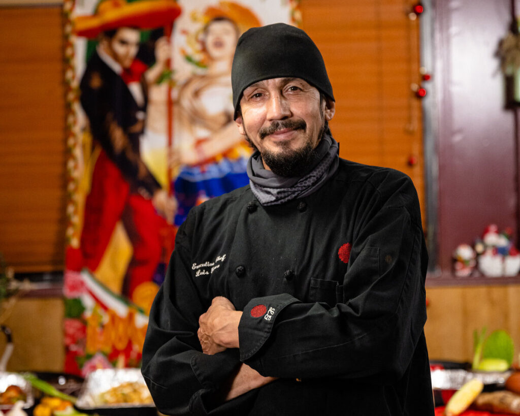 A man wearing a black beanie, grey scarf and black chef's coat smiles and poses for a photo with his arms crossed over his chest. 