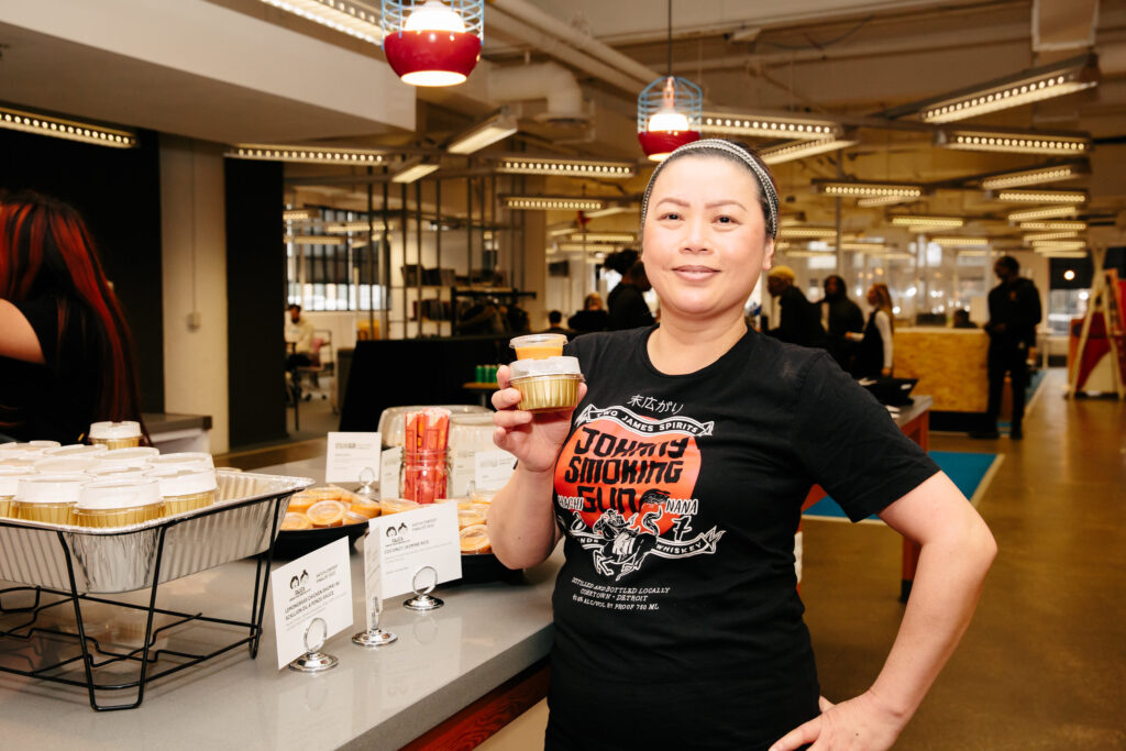 A woman wearing a black T-shirt with white, black and orange text and graphics smiles and holds little to-go containers of food text to her table setup of dumplings in to-go containers