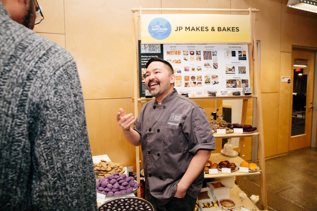 A man wearing a grey chef's jacket and black pants talks with another man. He's surrounded by various pastries on displays.