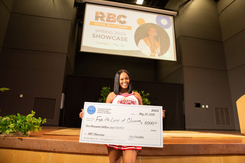 A woman stands in front of a stage in an auditorium. She is smiling and holding a large check for $5,000 from TechTown that she won for her business, For the Love of Cheesecake, at the Spring 2023 Retail Boot Camp Showcase.