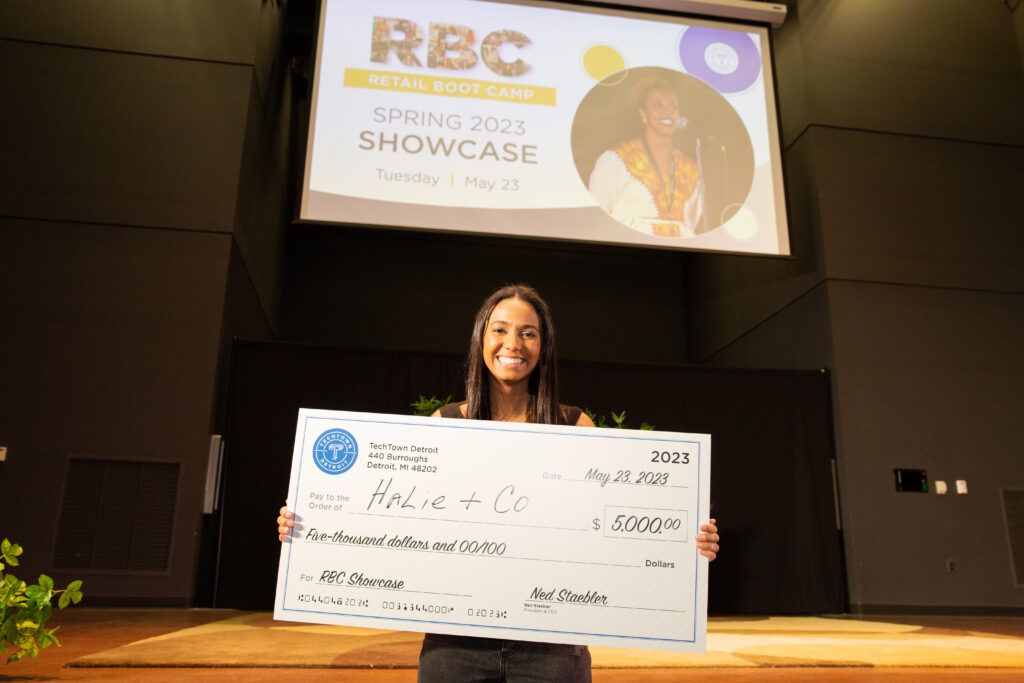 A woman stands in front of a stage in an auditorium. She is smiling and holding a large check for $5,000 from TechTown that she won for her business, Halie & Co.