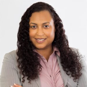 A woman of color smiles for a headshot photo. She is wearing a light grey blazer and mauve button-up shirt.