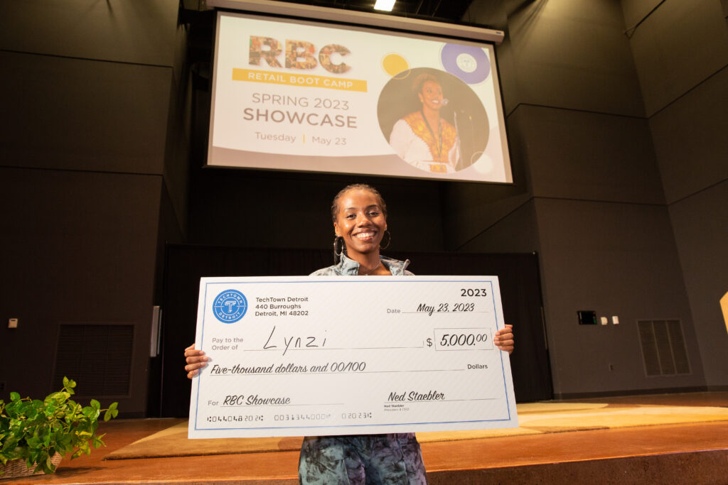 A woman stands in front of a stage in an auditorium. She is smiling and holding a large check for $5,000 from TechTown that she won for her business, Lynzi, at the Spring 2023 Retail Boot Camp Showcase.