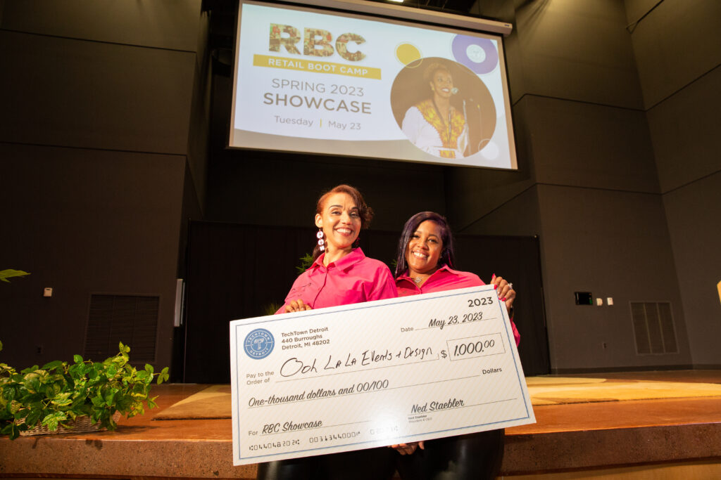 Two women stand in front of a stage in an auditorium. They are smiling and holding a large check for $1,000 from TechTown that she won for their business, Ooh La La Events & Design.