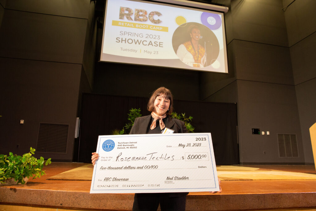A woman stands in front of a stage in an auditorium. She is smiling and holding a large check for $5,000 from TechTown that she won for her business, Rosemarine Textiles.