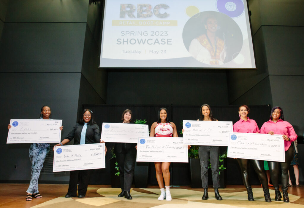 Seven women stand on a stage in an auditorium. They are each smiling and holding a large check from TechTown that they won for their businesses at the Spring 2023 Retail Boot Camp (RBC) Showcase. Behind them is a large screen hanging from the ceiling with a slide that says "RBC Retail Boot Camp" Spring 2023 Showcase Tuesday May 23"