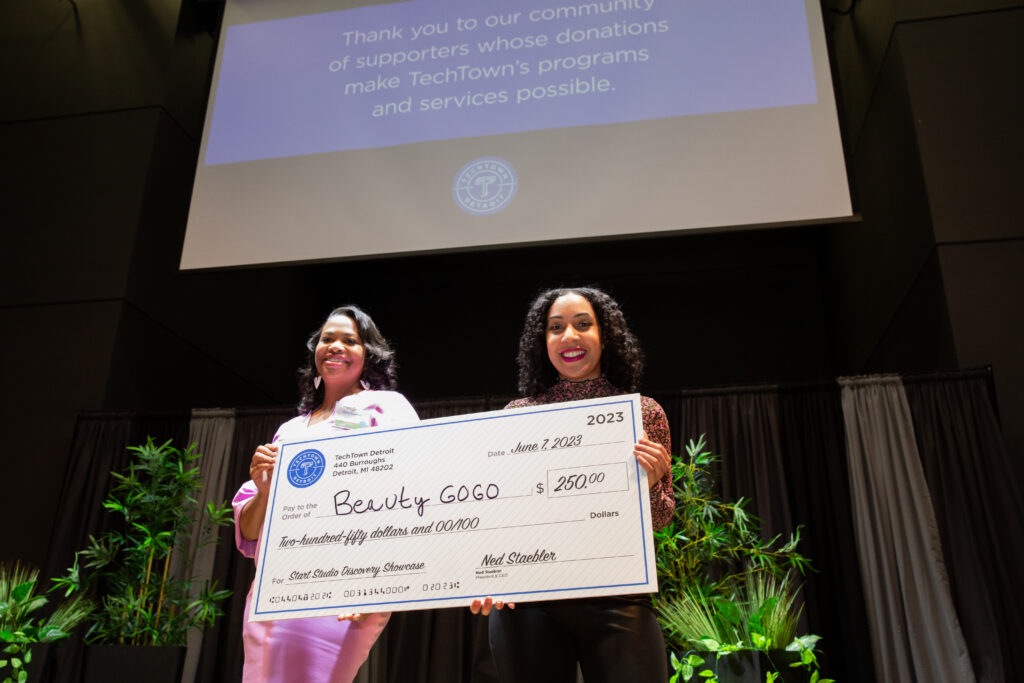 Two women stand on a stage and smile while holding a large check for $250 from TechTown Detroit, which they were awarded for their business, Beauty GoGO. Behind them is a screen hanging from the ceiling, presenting a slide that says "Thank you to our community of supporters whose donations make TechTown's programs and services possible."