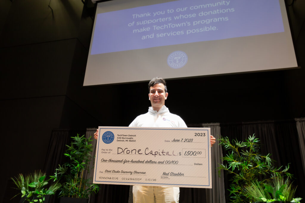 A man stands on a stage and smiles while holding a large check for $1,500 from TechTown Detroit, which he was awarded for his business, Drone Capital. Behind him is a screen hanging from the ceiling, presenting a slide that says "Thank you to our community of supporters whose donations make TechTown's programs and services possible."