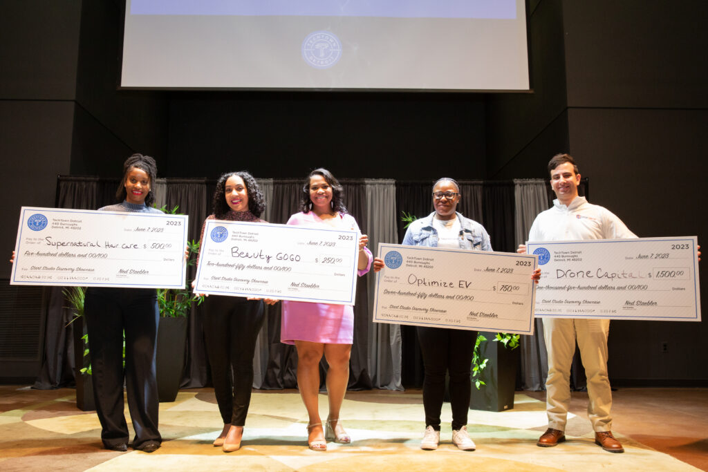A group of five women and men stand on a stage in an auditorium. Each of them are smiling and holding large checks from TechTown Detroit that they were each awarded at the Spring 2023 Start Studio Discovery Showcase