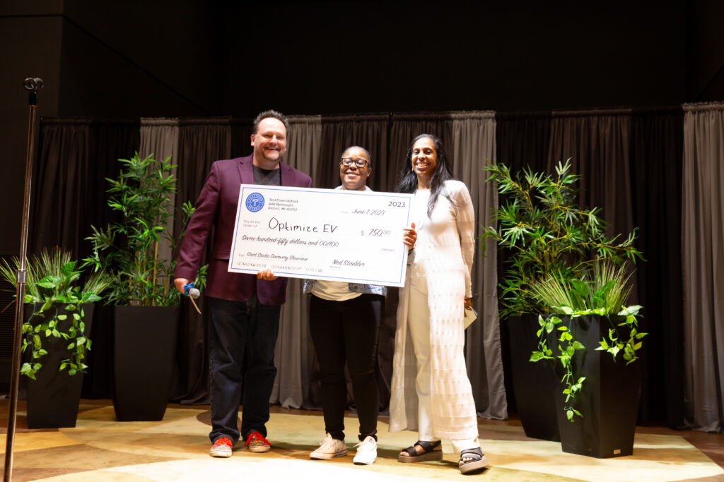 A woman stands on a stage and smiles while holding a large check for $750 from TechTown Detroit, which she was awarded for her business, OptimizeEV. Another woman is smiling and standing to her left and a man is smiling and standing to her right.