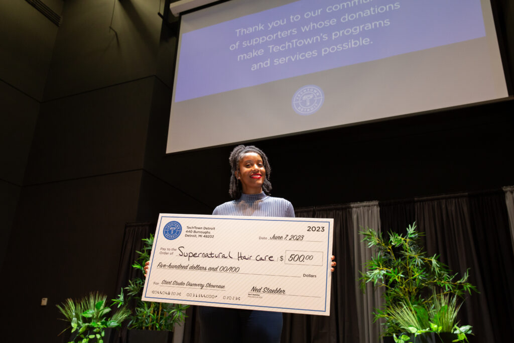 A woman stands on a stage and smiles while holding a large check for $500 from TechTown Detroit, which she was awarded for her business, Supernatural Hair Care. Behind her is a screen hanging from the ceiling, presenting a slide that says "Thank you to our community of supporters whose donations make TechTown's programs and services possible."