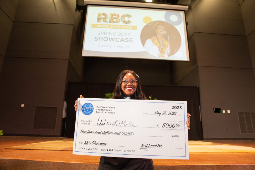 A woman stands in front of a stage in an auditorium. She is smiling and holding a large check for $5,000 from TechTown that she won for her business, U Drink I Make, at the Spring 2023 Retail Boot Camp Showcase.