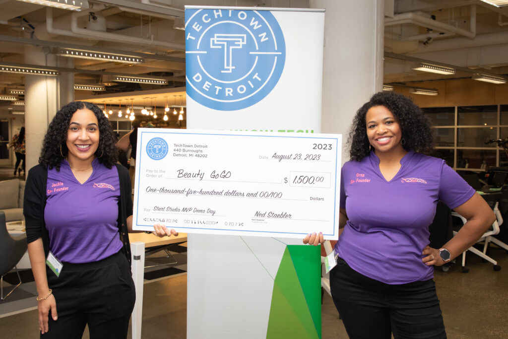 Two women of color hold up a large check for $1,500, for their business, Beauty GoGO