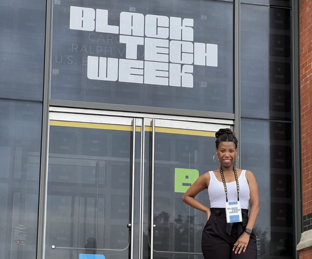 A woman stands outside of a building and smiles for a photograph. Behind her is a large set of doors and a window with a white vinyl decal saying "Black Tech Week."