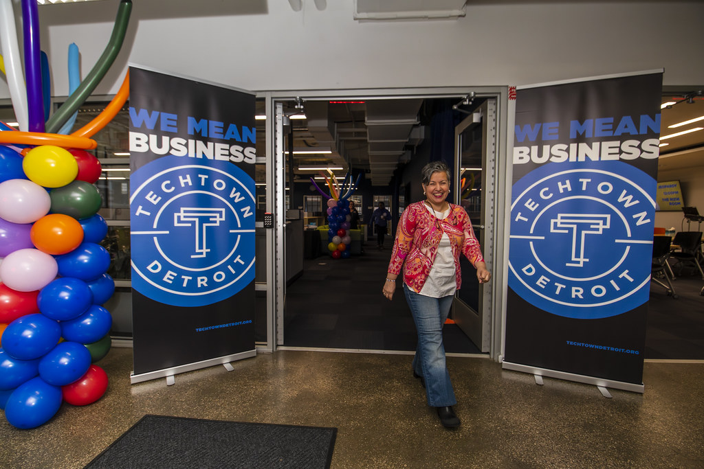 A woman smiles as she walks out two double doors. On each side of the doors are large posters on stands that say "We Mean Business" followed by TechTown's blue-and-white logo. Next to the flyer on the far left side is a large, multicolor balloon display