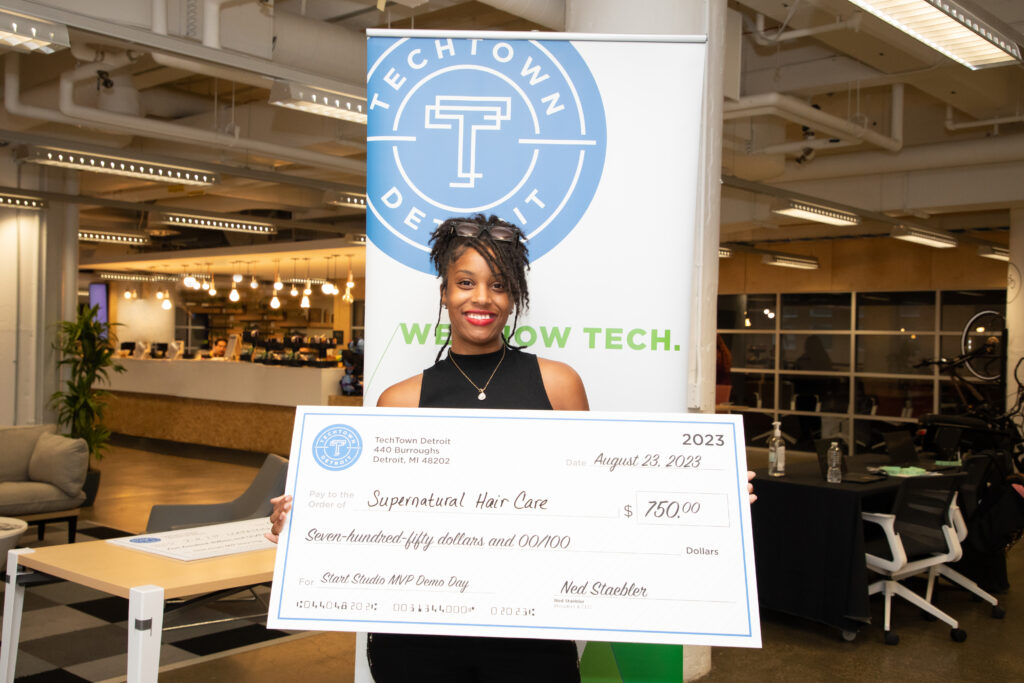 A woman of color holds a large check for $750, for her business, Supernatural Hair Care