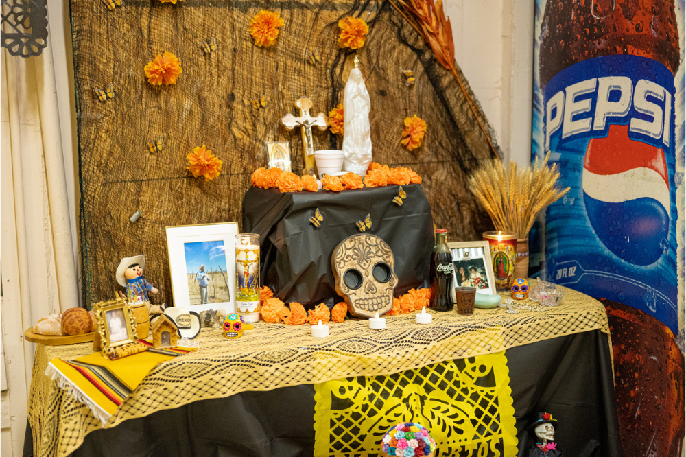 An altar featuring ofrendas for Day of the Dead. The display includes a wooden skull, flowers, a small cross figurine, an angel figurine, small candles, and framed photos