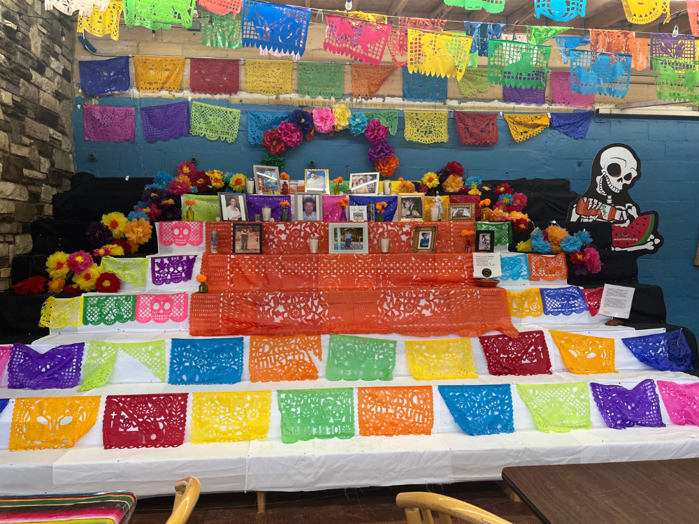 A display of ofrenda for Day of the Dead at La Jalisciense. The display includes multicolor paper decorations, photos, flowers and candles
