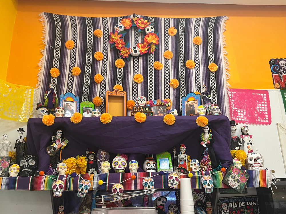 A display of ofrenda for Day of the Dead at Mangonadas del Barrio. The display includes multicolor flowers, decorated skulls, framed photos and candles