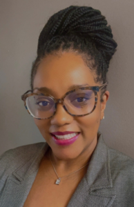 A photo of a woman of color. She is smiling and wearing a gray blazer and brown tortoise glasses