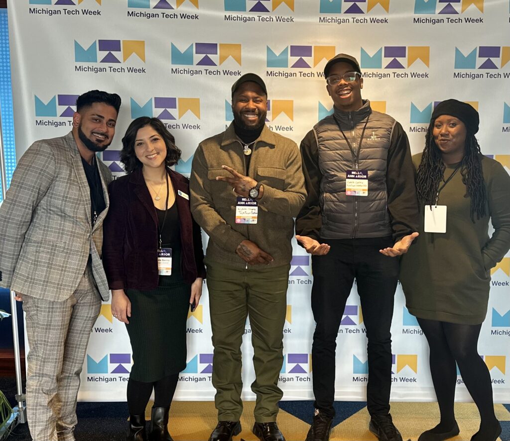 Five members of TechTown Detroit's Tech-based Programs team gather and smiling in front of a step-and-repeat banner at Michigan Tech Week