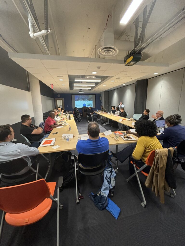 A group of people sit at long desks, round-table style, for a workshop held in a conference room. They're all looking at the front of the room, where two people are talking in front of a projector. 