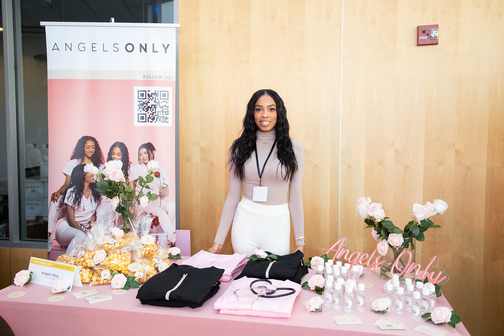 A woman smiles and stands behind a table display of medical scrubs and accessories. Beside her is a banner featuring her business Angels Only