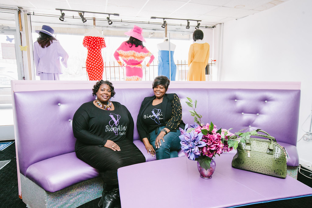 Two women sit inside a clothing store on a purple couch, near a purple table with flowers and a purse sitting on it. Behind them are mannequins set up on window display