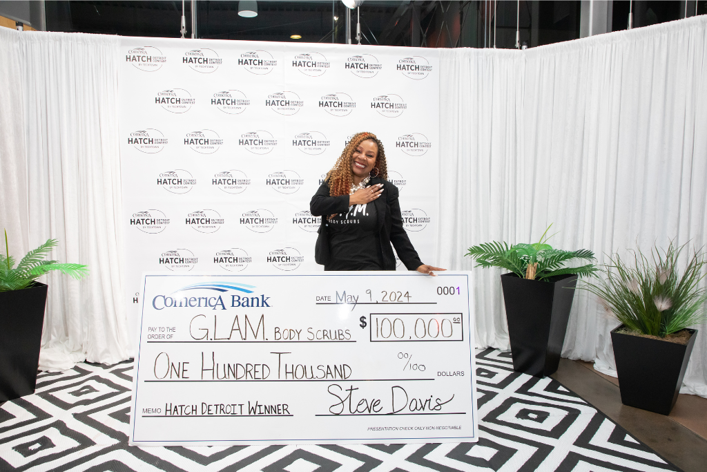 A woman smiles while holding her right hand over her heart. With her left hand, she holds a large check for $100,000 from Comerica Bank for her business, G.L.A.M. Body Scrubs, the winner of the 2024 Comerica Hatch Detroit Contest by TechTown