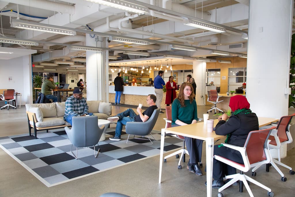 A photo of coworking office. People are sitting at tables, on chairs and at couches. Towards the back of the space is a front desk that people are sitting and standing around.