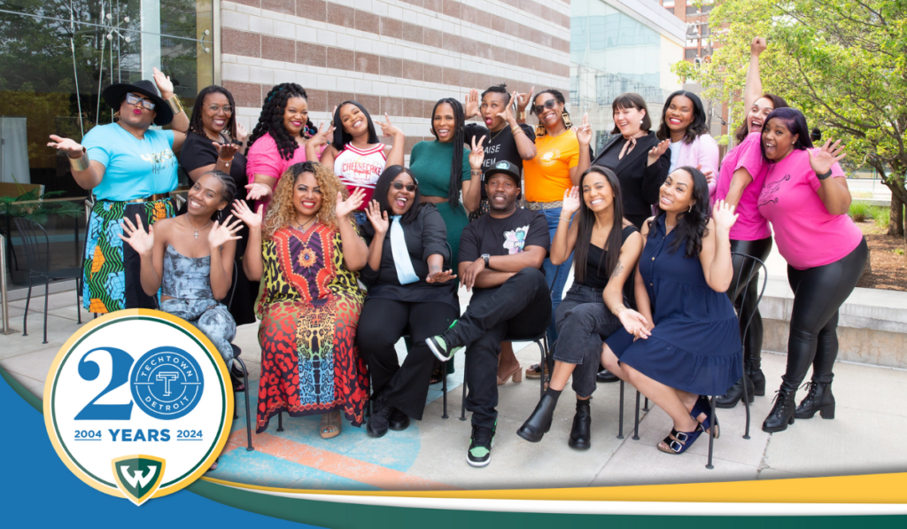 A large group photo of TechTown alumni smiling and being excited. At the bottom of the photo, there is a horizontal yellow, green and blue ribbon and TechTown's 20th anniversary logo. 