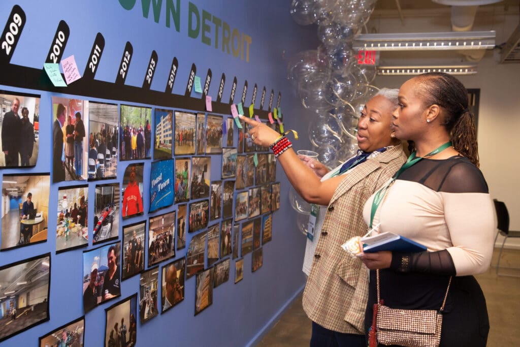 Two individuals look at rows of photos on a wall, organized in a timeline fashion