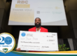 A photo of Scott Boateng holding a large check for $1,000 from TechTown Detroit for his business, Preva Body. He is standing on an auditorium stage. Behind him is a projector screen displaying a slide for TechTown Detroit's Retail Boot Camp: E-commerce & Home-based Edition program and thanking its sponsors