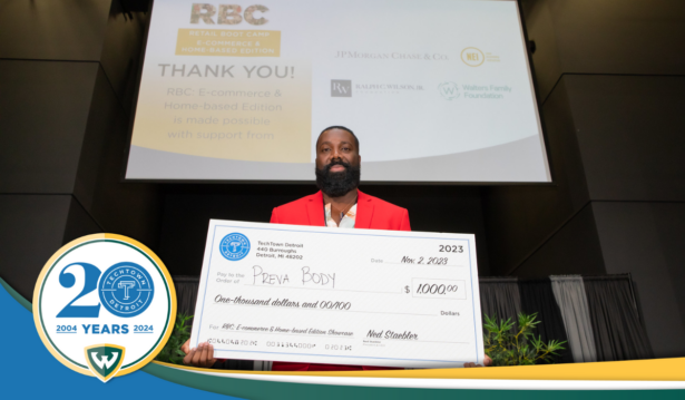 A photo of Scott Boateng holding a large check for $1,000 from TechTown Detroit for his business, Preva Body. He is standing on an auditorium stage. Behind him is a projector screen displaying a slide for TechTown Detroit's Retail Boot Camp: E-commerce & Home-based Edition program and thanking its sponsors