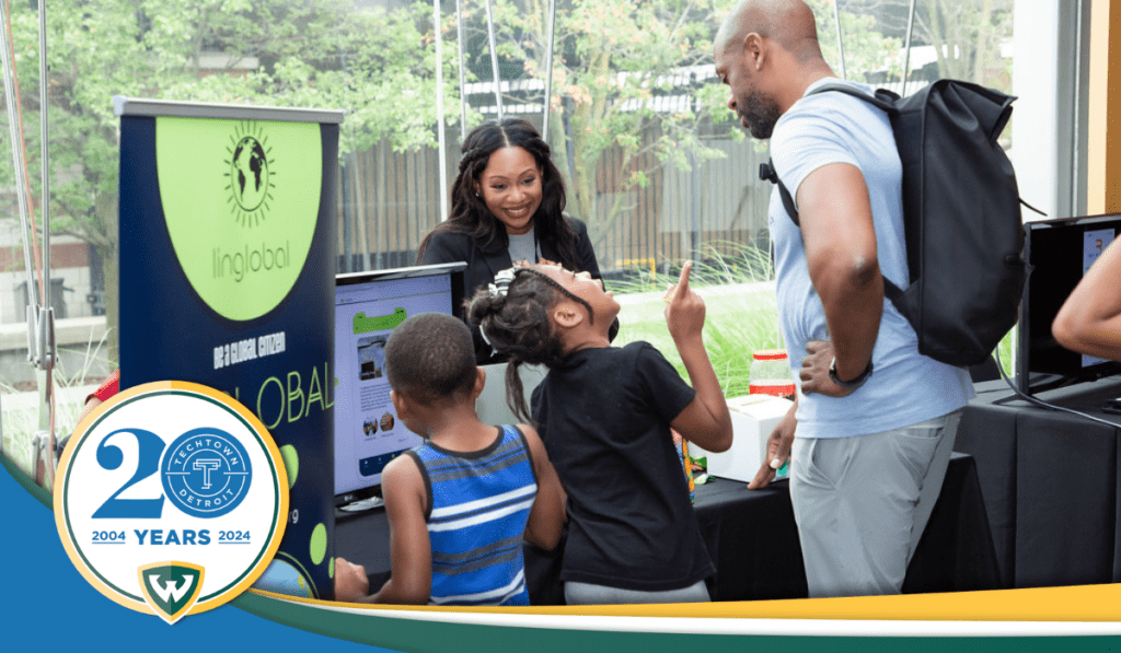 A photo of tech founder Ernestine Lyons, creator of Linglobal, stands behind a table display for her startup. In front of it are two kids and adult looking at the items on the table. Overlapping the photo is the TechTown Detroit 20th anniversary logo