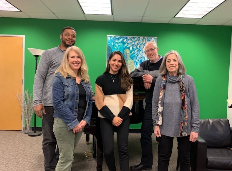 A group of five people smile and pose for a photograph in front of a piano. They're inside a room with a green wall with a painting on it, a floor lamp and a sofa chair.