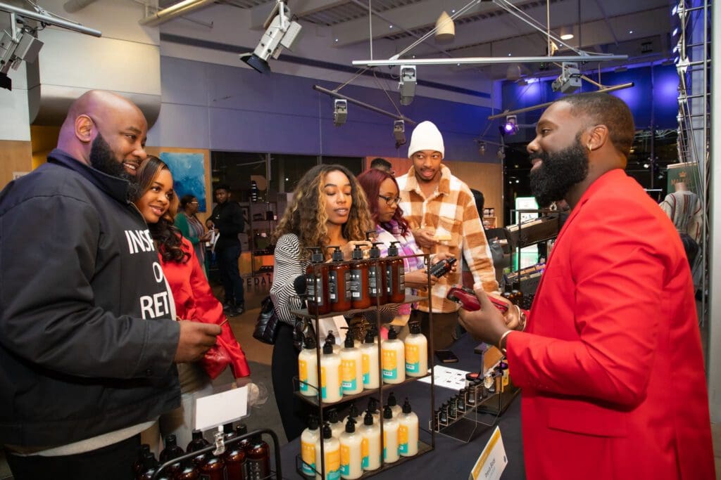 Scott Boateng stands behind a table display featuring items from his business Preva Body. He smiles as he interacts with several people standing around the display.