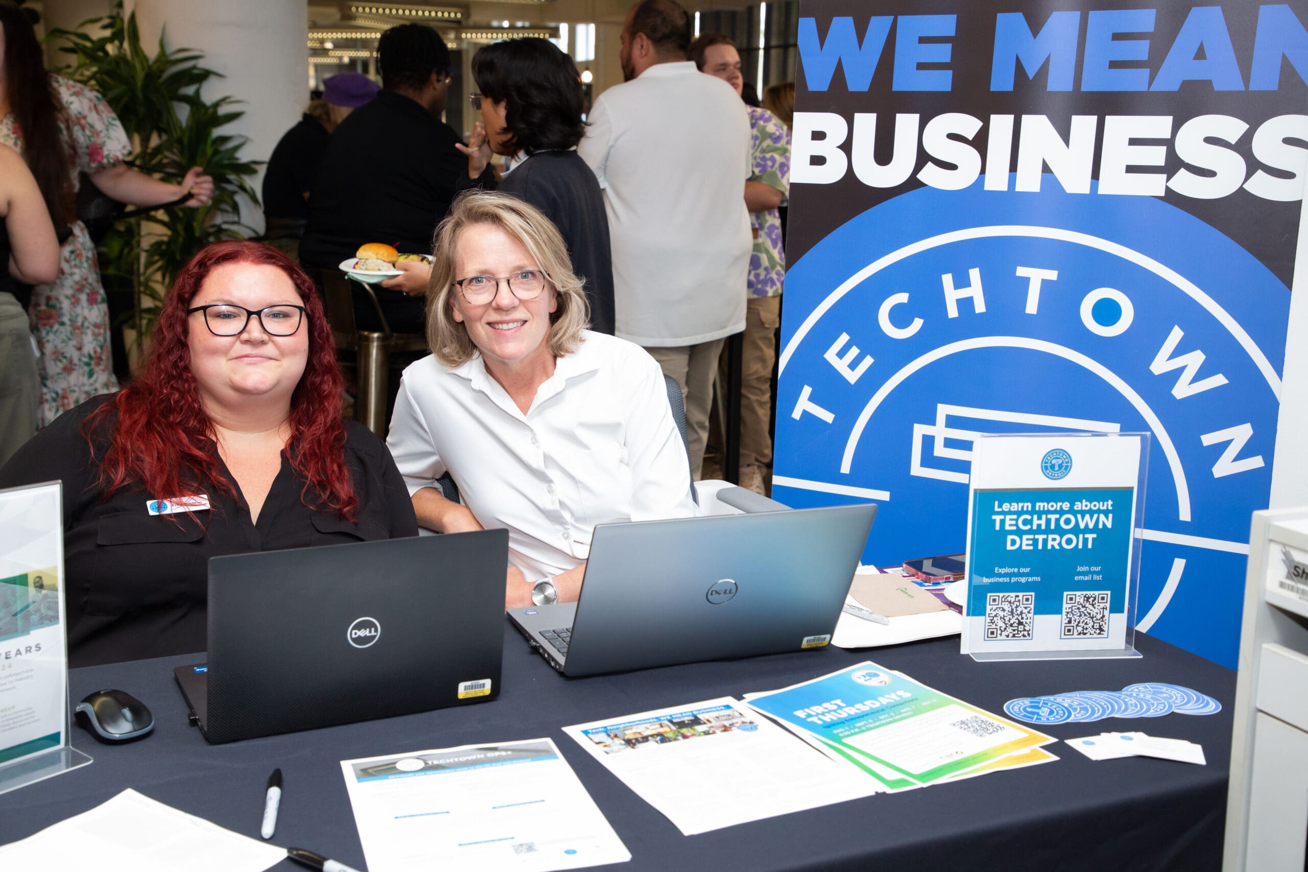 Two individuals seated at a promotional booth with laptops, in front of a banner. Flyers and informational materials are visible on the table.