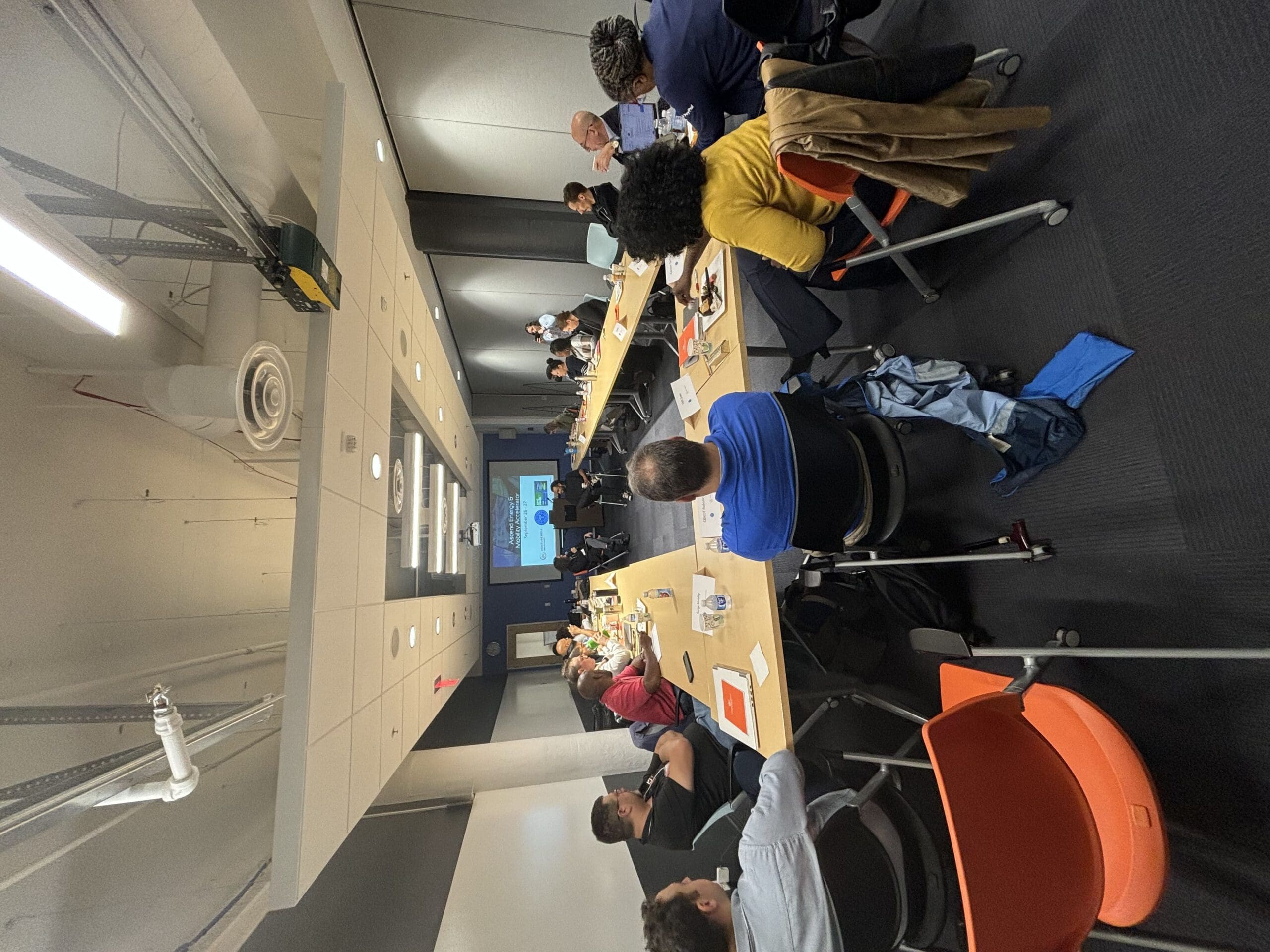 A group of people sit at long desks, round-table style, for a workshop held in a conference room. They're all looking at the front of the room, where two people are talking in front of a projector. 