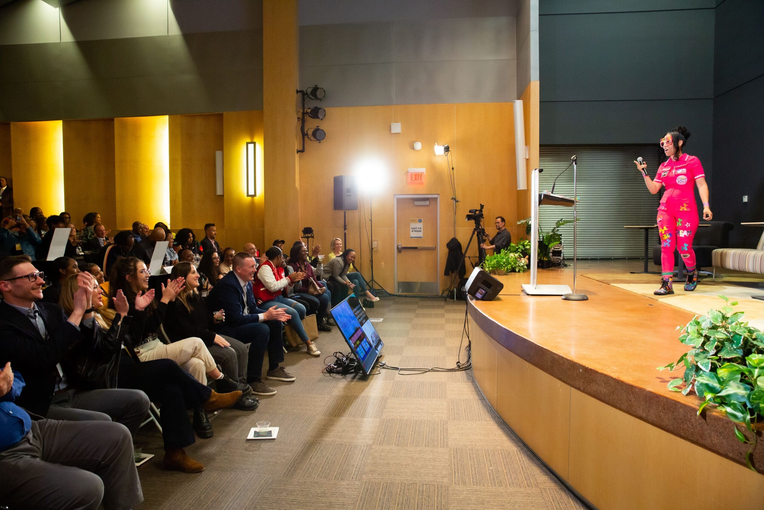A woman wearing a pink jumpsuit and glasses stands on stage and holds a microphone. She's talking to a live audience in an auditorium. The audience is clapping and smiling.