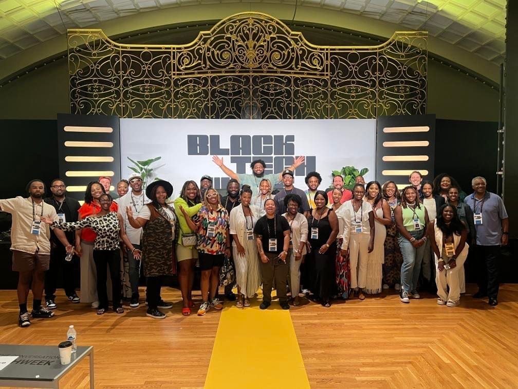 A group of people smile and pose for a photograph in a large room. Behind them is a screen that says "Black Tech Week"