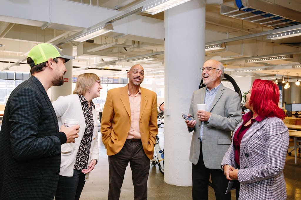 A group of two women and three men stand in an open office space talking and laughing