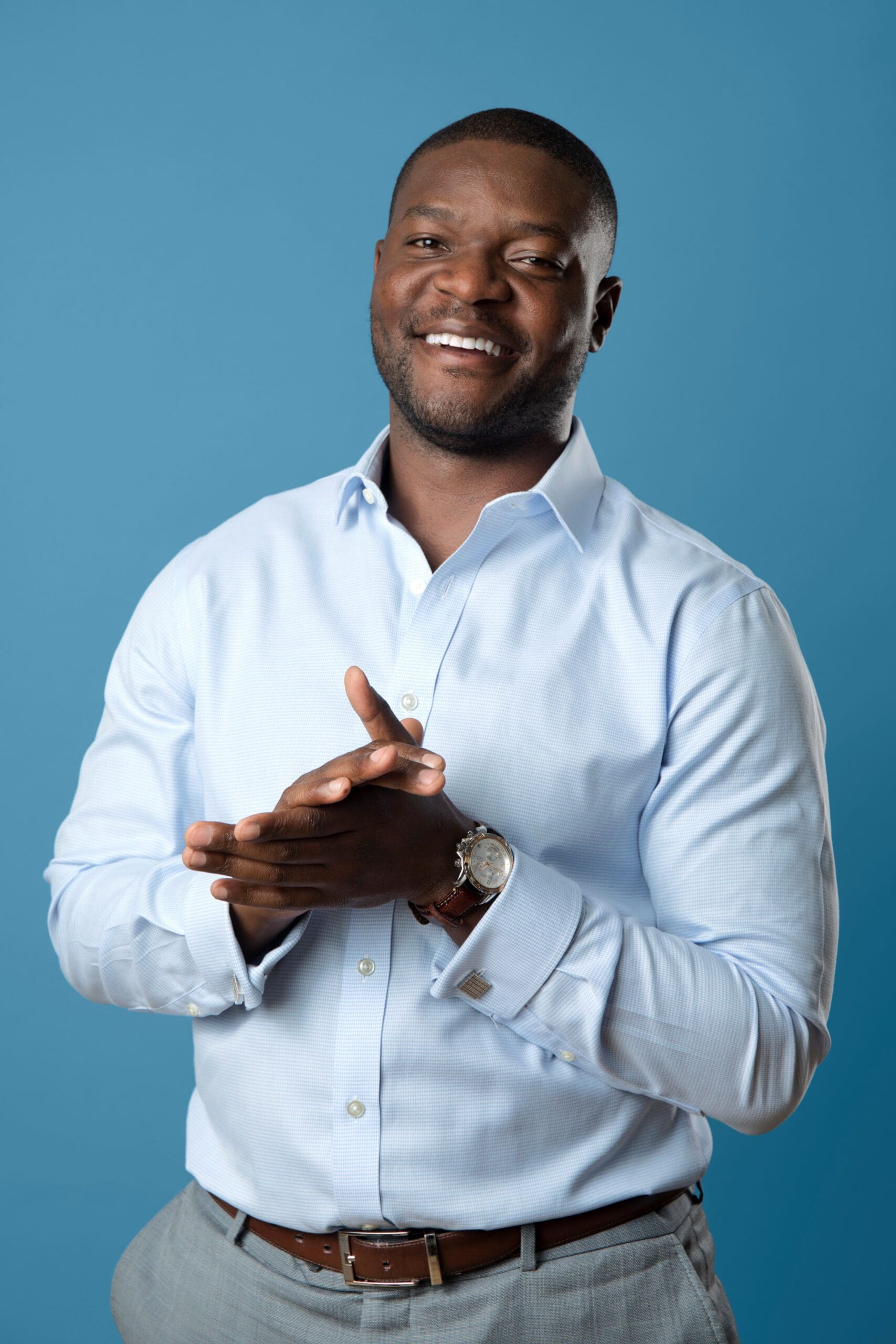 A man of color wearing a pale blue, button-down dress shirt a brown belt, and light grey pants. He's smiling and hand his hands clapped together. 