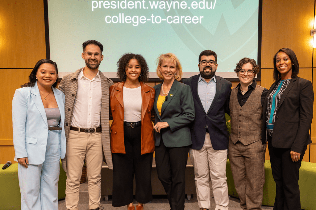 A group of seven people smile for a photograph while standing on the stage of an auditorium