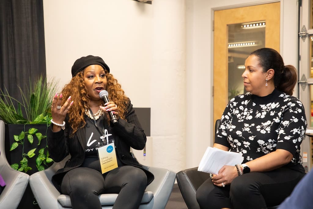 A woman speaks into a microphone, sitting next to another woman who is looking at her as she talks