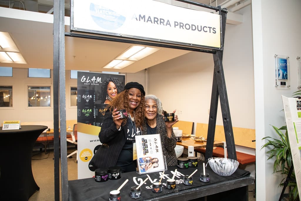 Two women smile while holding up products from the organic skincare line G.L.A.M. Body Scrubs, by Amarra Products. They stand behind a table display featuring products from G.L.A.M.